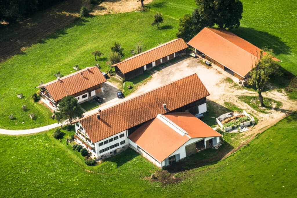 Vue aérienne d'un corps de ferme traditionnel avec toits tuilés rouges, maisons, granges et champs verts ensoleillés.