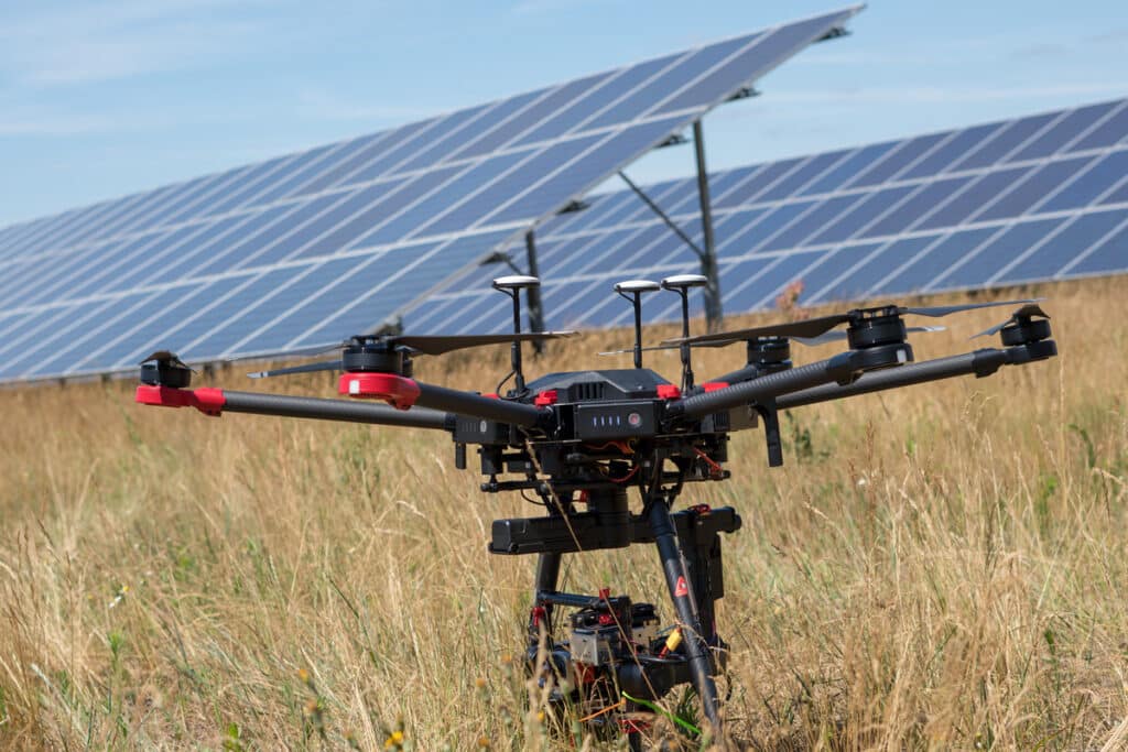 Grand drone professionnel noir et rouge posé dans un champ d'herbe sèche, avec des rangées de panneaux solaires en arrière-plan.