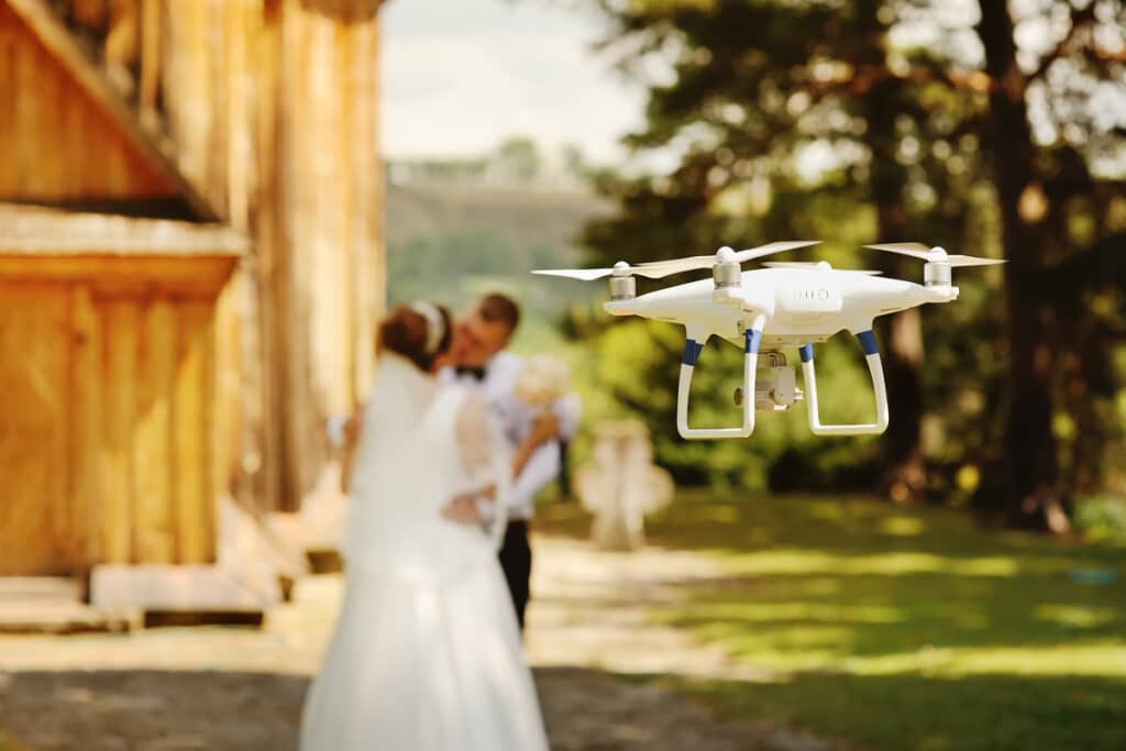 Drone blanc filme un couple de mariés s'embrassant en arrière-plan flou, dans un cadre extérieur verdoyant près d'un bâtiment en bois.