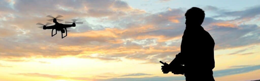 Silhouette d'un pilote contrôlant un drone en vol au-dessus d'un ciel orangé et bleu au coucher du soleil.